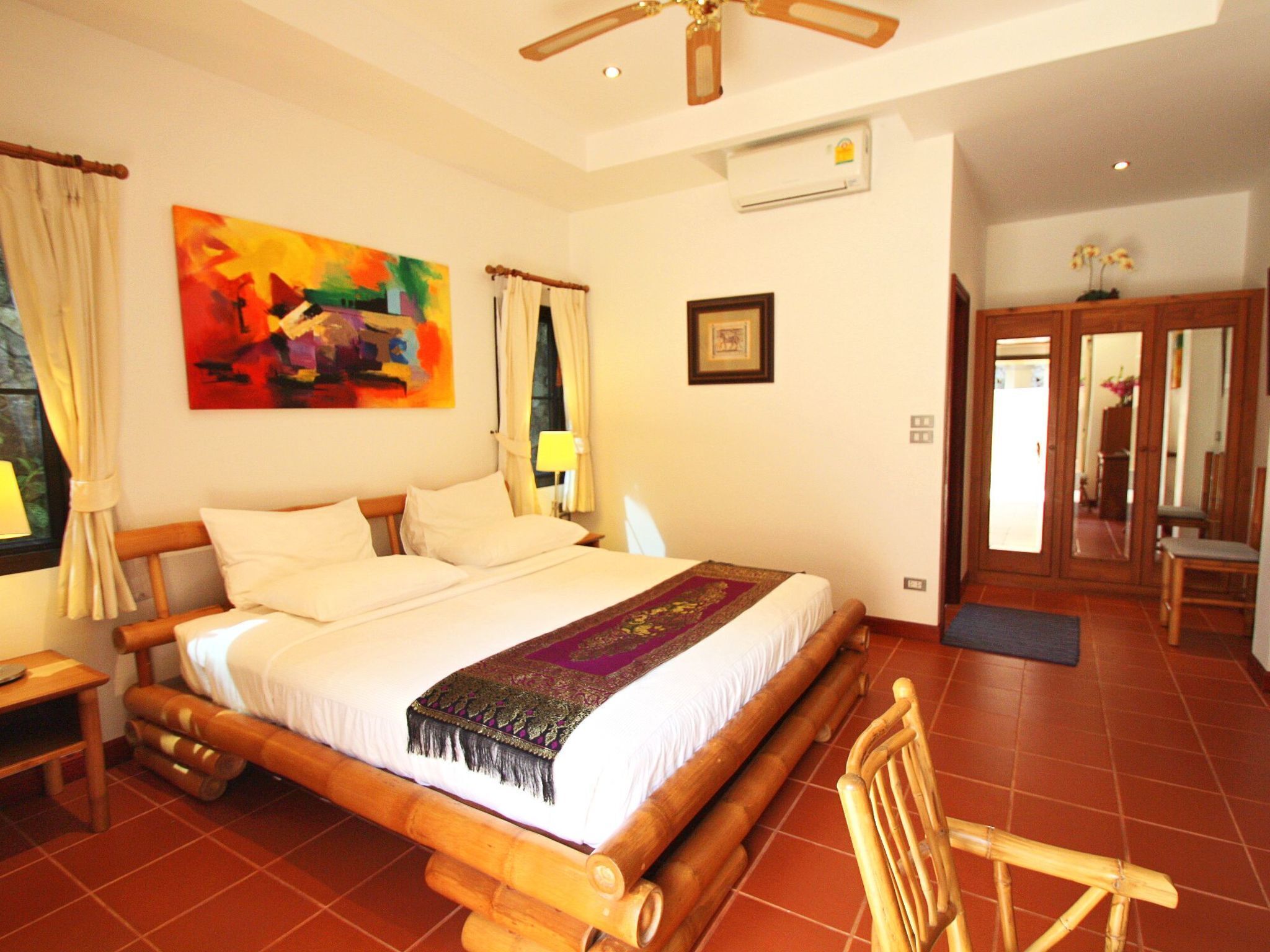 A room with a large bed, wooden headboard, a ceiling fan, a painting on the wall, and a door leading to another room.