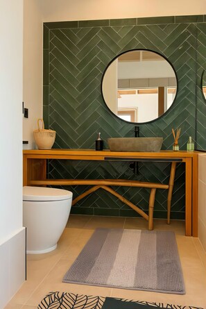 A modern bathroom with a herringbone-patterned green wall, a round mirror, a wooden vanity, a white toilet, and a grey rug.