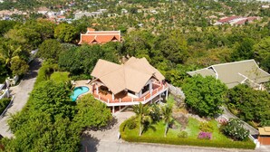 Vista aérea de un área residencial con una casa destacada que cuenta con una gran piscina, rodeada de exuberante vegetación y una vista despejada del océano.