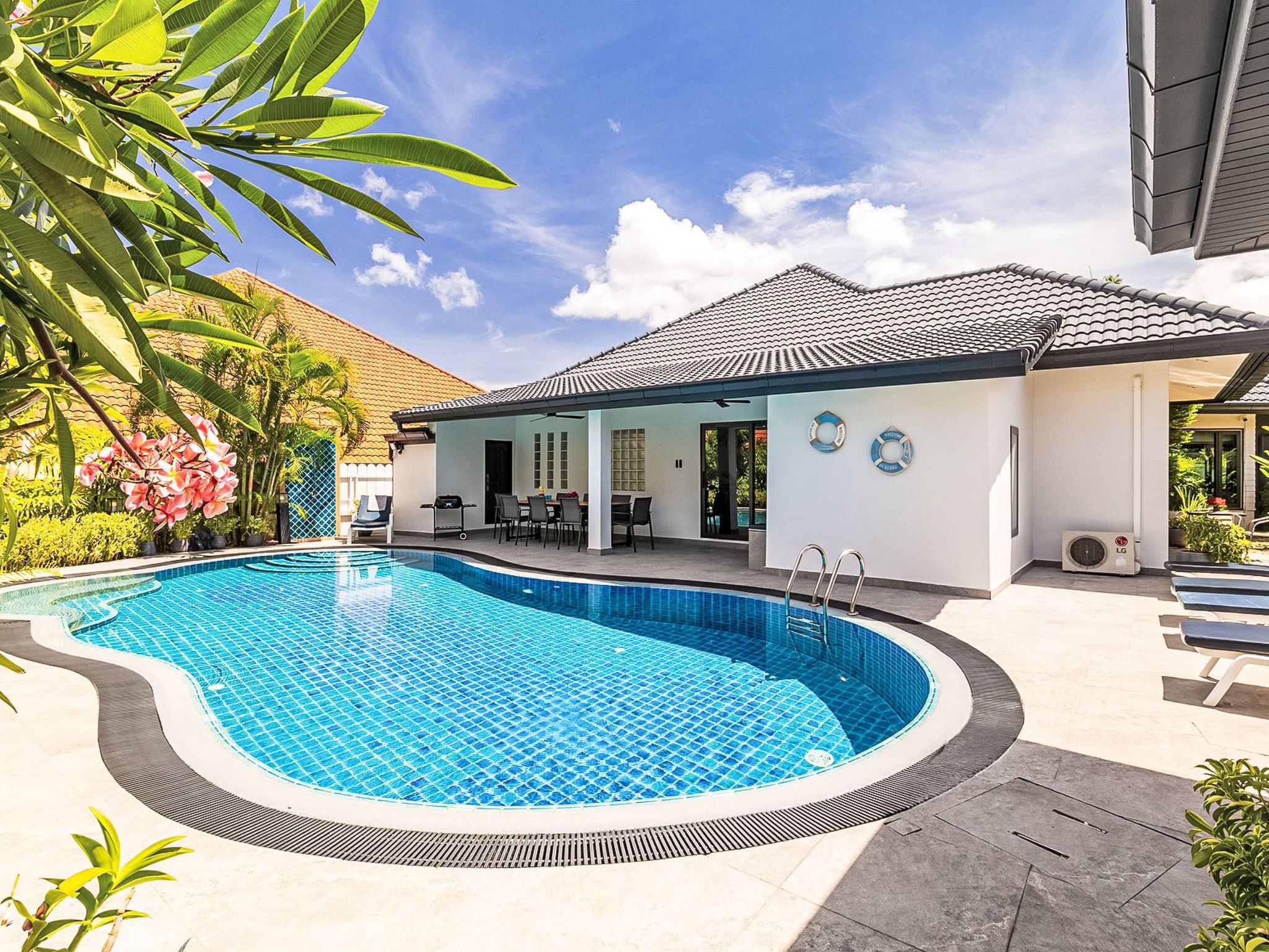 Ein Pool mit Fliesenrand, eine überdachte Terrasse mit Sitzgelegenheiten und ein Gebäude mit einem gefliesen Dach.
