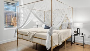 Una cama con dosel y cuatro columnas, con cubrecama y almohadas blancas, una mesita de noche con lámpara y una ventana con persianas.