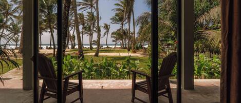 A room with a large glass door leading to a balcony with a view of a beach, palm trees, and a lawn.