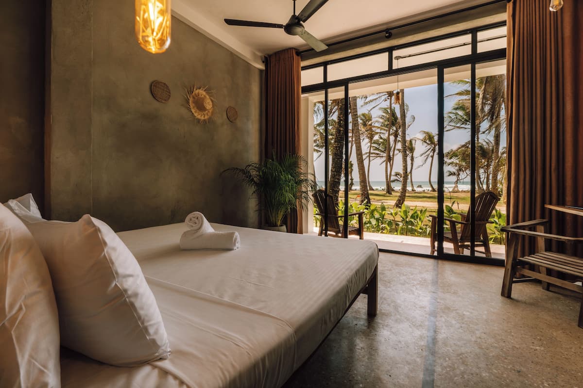 A bedroom with a large bed, a ceiling fan, a view of the beach, and a balcony with chairs.