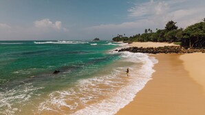 A beach with clear turquoise water, a sandy shore, and a rocky outcrop in the distance. - Surfescape Sri Lanka (Ahangama)