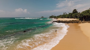 A beach with clear turquoise water, a sandy shore, and a rocky outcrop in the distance. - Surfescape Sri Lanka (Ahangama)