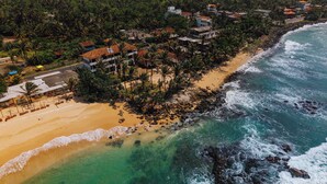 An aerial view of a coastal area with a beach, ocean, and residential buildings surrounded by palm trees. - Surfescape Sri Lanka (Ahangama)