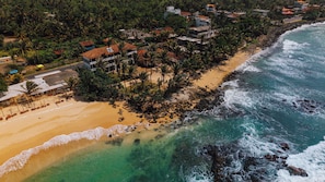 An aerial view of a coastal area with a beach, ocean, and residential buildings surrounded by palm trees. - Surfescape Sri Lanka (Ahangama)