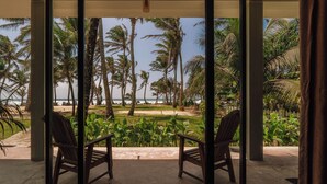 A room with a large glass door leading to a balcony with a view of a beach, palm trees, and a lawn. - Surfescape Sri Lanka (Ahangama)
