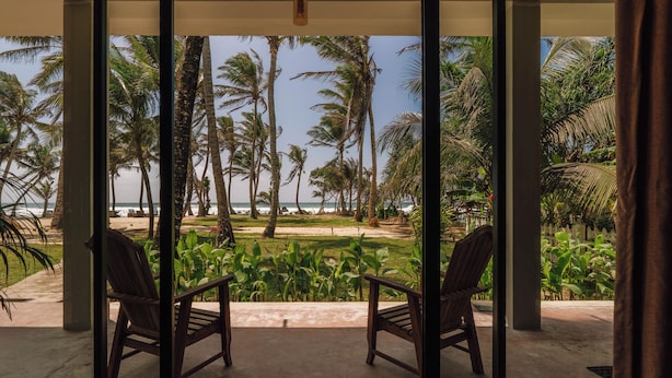 A room with a large glass door leading to a balcony with a view of a beach, palm trees, and a lawn. - Surfescape Sri Lanka (Ahangama)