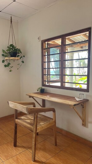A room with a wooden desk, a chair, a hanging plant, and a window with a view of greenery. - Surfescape Sri Lanka (Ahangama)