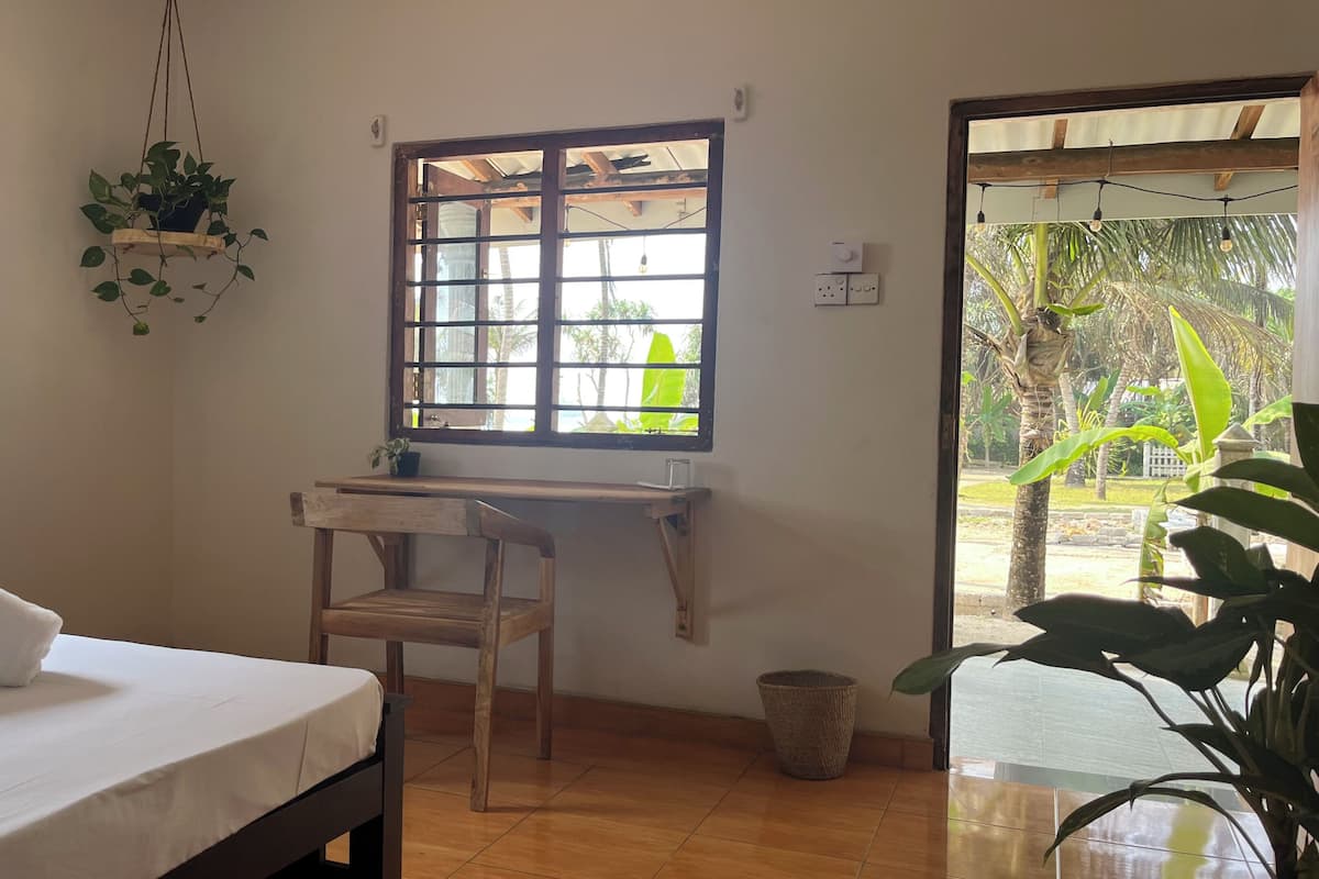 A room with a bed, a desk, a chair, a hanging plant, a window, and a view of the outdoors.