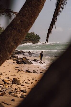A beach scene with a person standing near the water, rocks, and a palm tree. - Surfescape Sri Lanka (Ahangama)