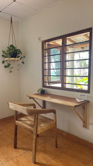 Ein Zimmer mit Schreibtisch aus Holz, einem Stuhl, einer hängenden Pflanze und einem Fenster mit Blick ins Grüne.