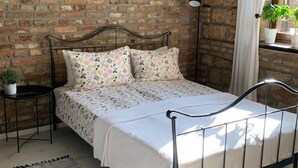 Un lit avec un cadre en métal, une couvre-lit blanche et des coussins à motifs floraux, dans une pièce dont les murs sont en briques apparentes et dont le plafond est en bois.