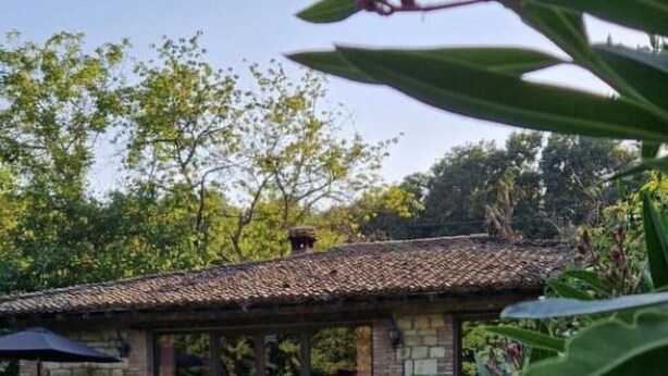 Une piscine avec une terrasse en pierre, entourée de verdure et un bâtiment en pierre doté d’un toit en tuiles.
