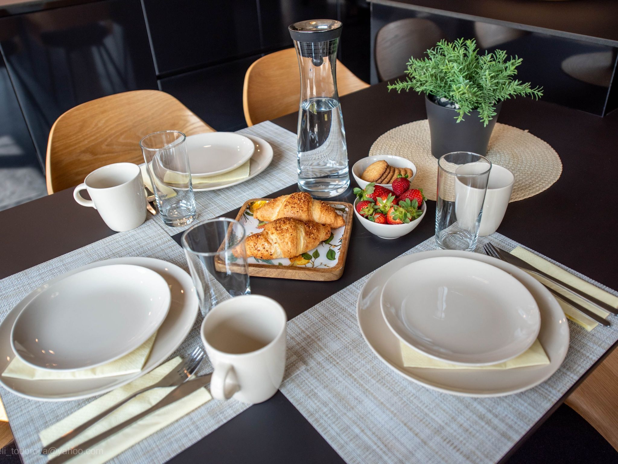 Een eettafel gedekt met borden, bestek, een karaf, glazen, een schaal met aardbeien, croissants en een bloempot.