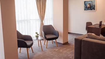 Ein modernes Hotelzimmer mit großem Fenster, einer Couch, zwei Sesseln und einem kleinen Tisch mit einem Laptop.
