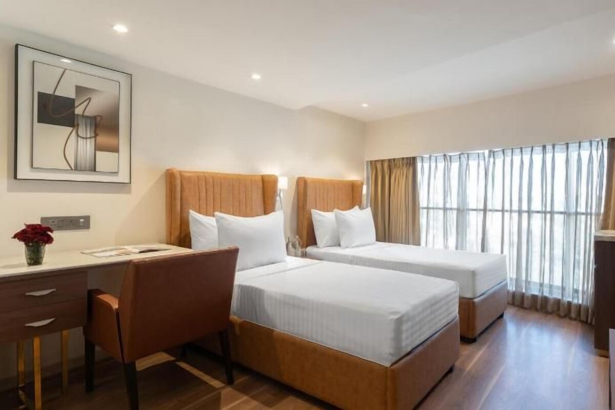 Ein Hotelzimmer mit zwei Betten, einem Schreibtisch, einem Stuhl, einer Kaffeemaschine und einem großen Fenster mit Vorhängen.