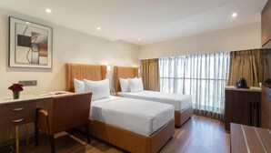 Ein Hotelzimmer mit zwei Betten, einem Schreibtisch, einem Stuhl, einer Kaffeemaschine und einem großen Fenster mit Vorhängen.
