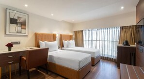 Ein Hotelzimmer mit zwei Betten, einem Schreibtisch, einem Stuhl, einer Kaffeemaschine und einem großen Fenster mit Vorhängen.
