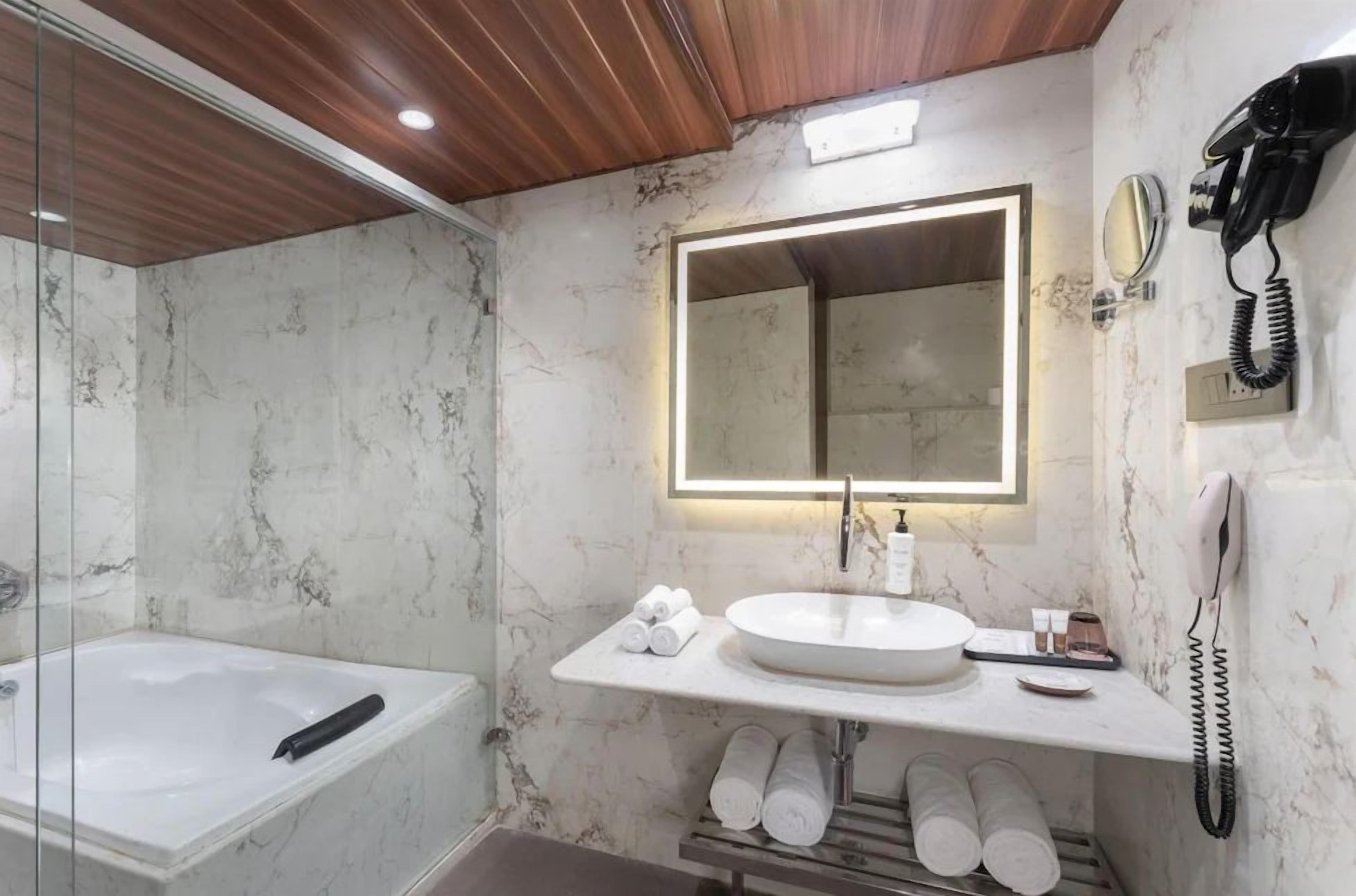 A modern bathroom with a marble wall, a large mirror, a sink, a bathtub, and a telephone.