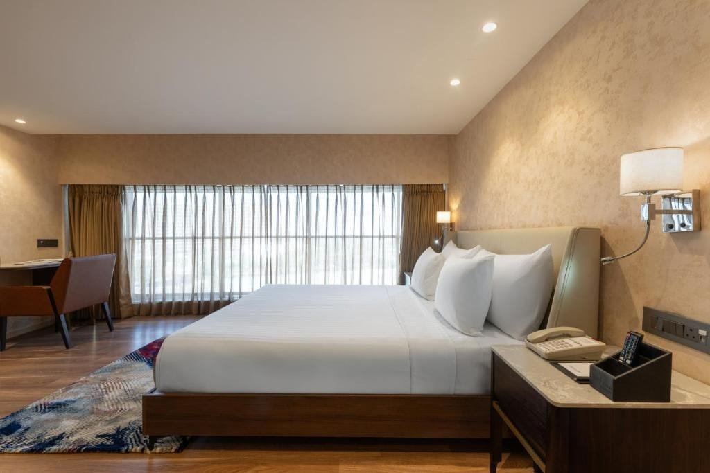 A hotel room with a large bed, a desk, a chair, a lamp, and a phone.