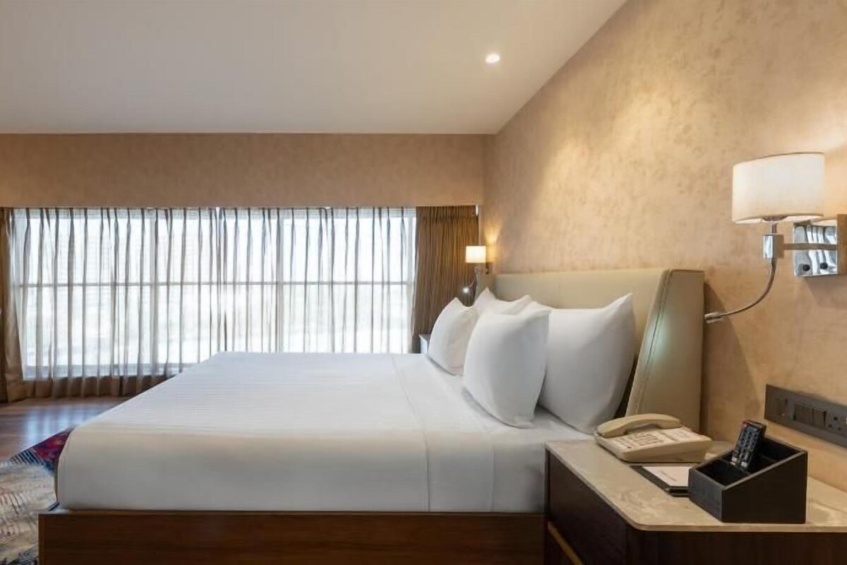 Ein Hotelzimmer mit einem großen Bett, einem Schreibtisch, einem Sessel, einem Nachttisch mit Telefon und einem Vorhangfenster.