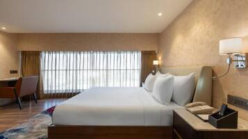 A hotel room with a large bed, a desk, a chair, a bedside table with a phone, and a window with curtains.