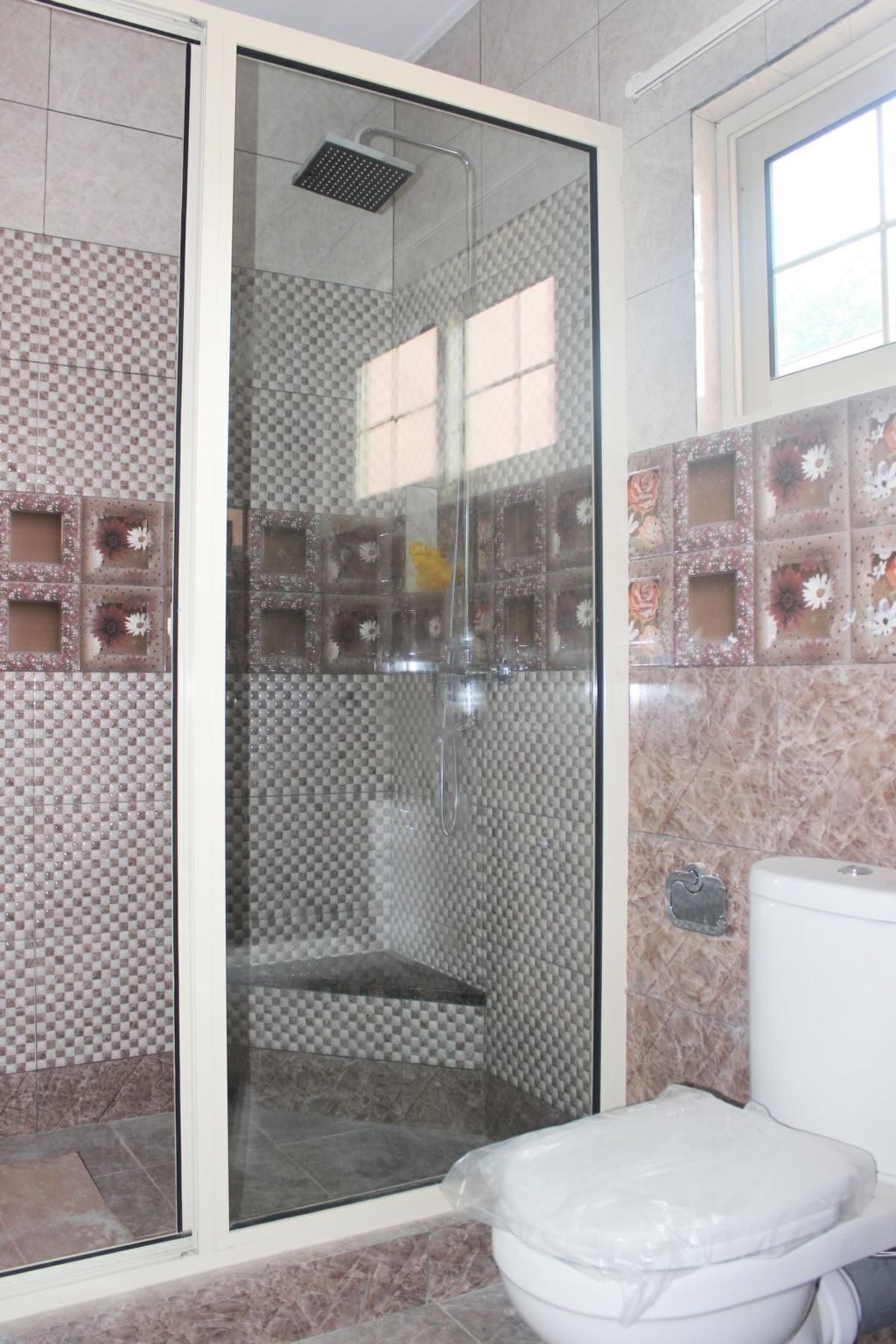 Kylpyhuone, jossa on lasiseinällä varustettu suihku, laatoitetut seinät ja wc.