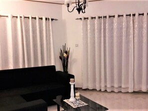 Un salon avec un canapé noir, une table basse en verre et des rideaux blancs.