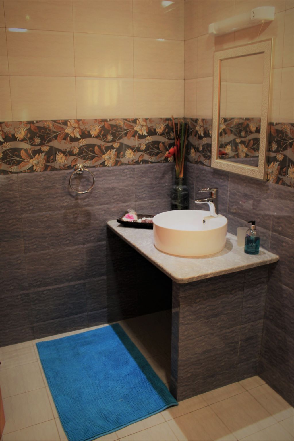 Ein Badezimmer mit einem runden Waschbecken, dunklen gemusterten Fliesen, einer blauen Badmatte und einem Spiegel über dem Waschbecken.