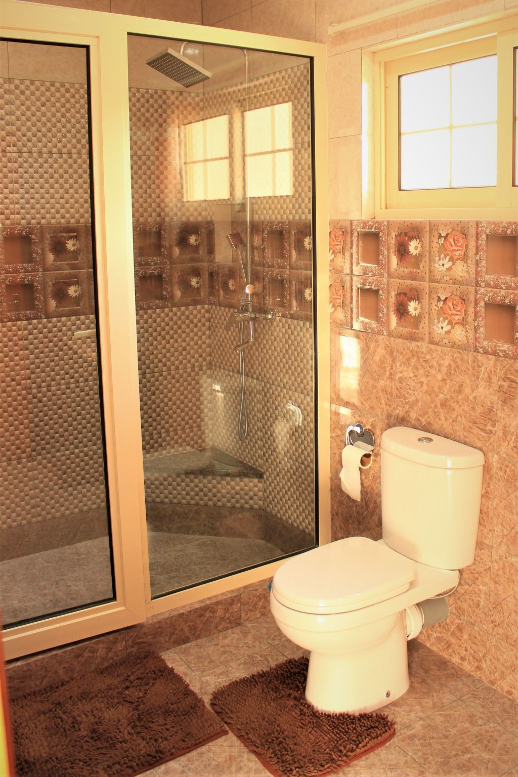 Et badeværelse med glasbrusekabine, toilet og et vindue med blomstermønstrede fliser.
