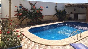 A swimming pool with a tiled deck, surrounded by a white wall with climbing plants and a wooden pergola. - Studio mit Pool, Garten und Dachterrasse by Interhome (Luxor)