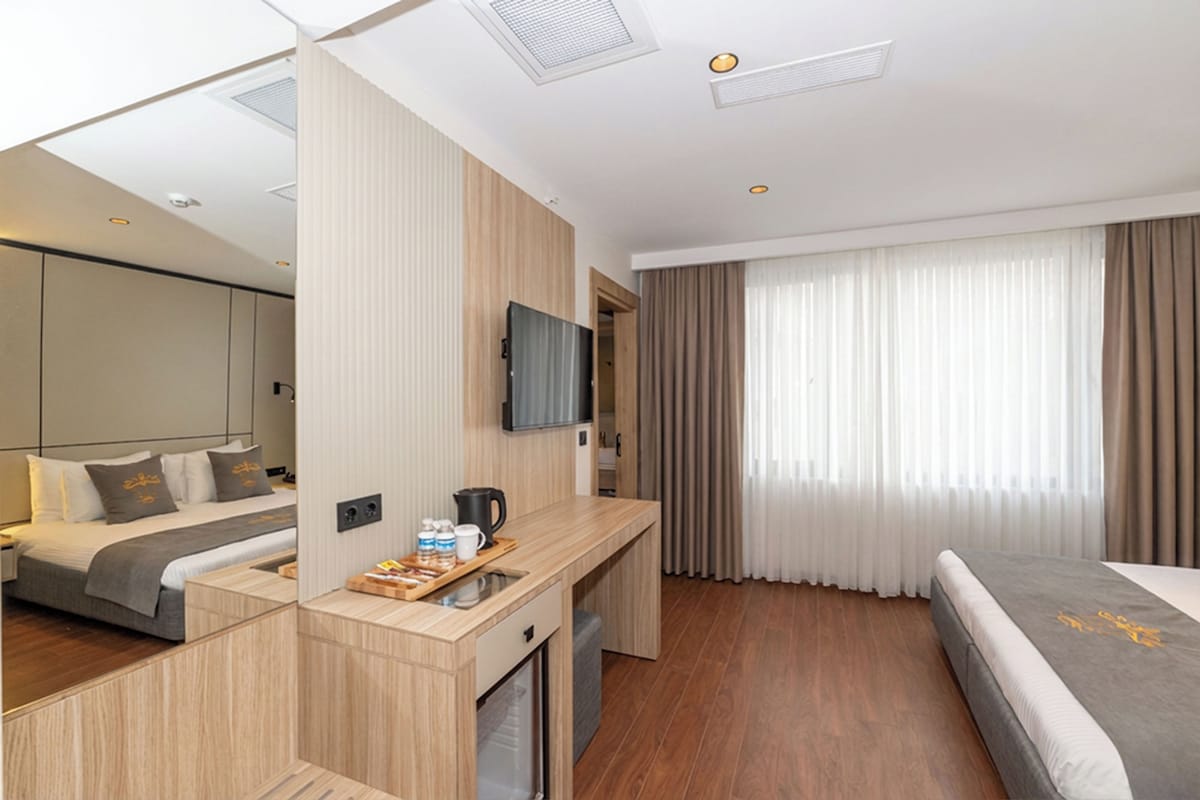 A modern hotel room with a bed, a desk with a television, a mirror, and a large window with curtains.