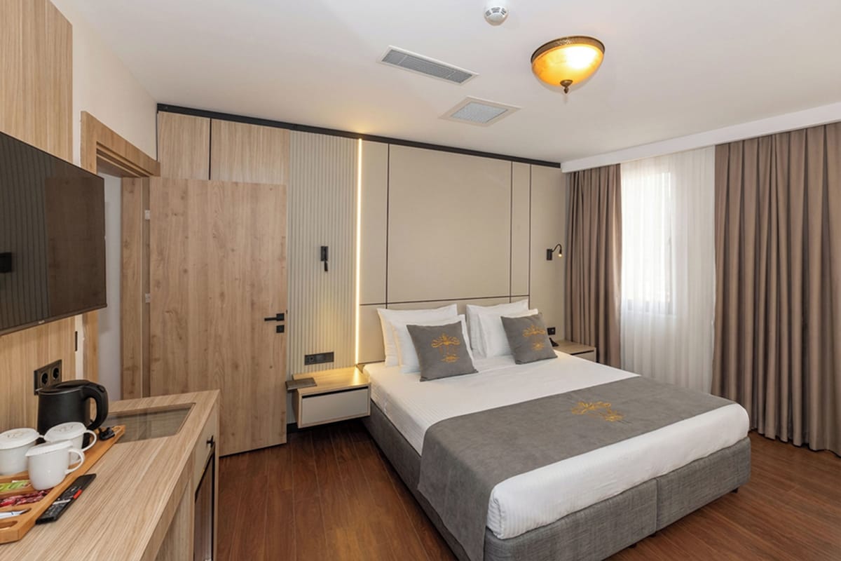 A modern hotel room with a large bed, wooden furniture, a flat-screen TV, and a bedside table with a kettle.