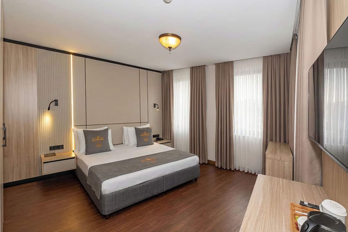 A modern hotel room with a large bed, wooden flooring, a desk with a lamp, and a large window with curtains.