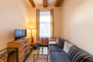 Un espace de vie compact comprenant un canapé, un téléviseur, une lampe, une petite table et une fenêtre avec des rideaux.