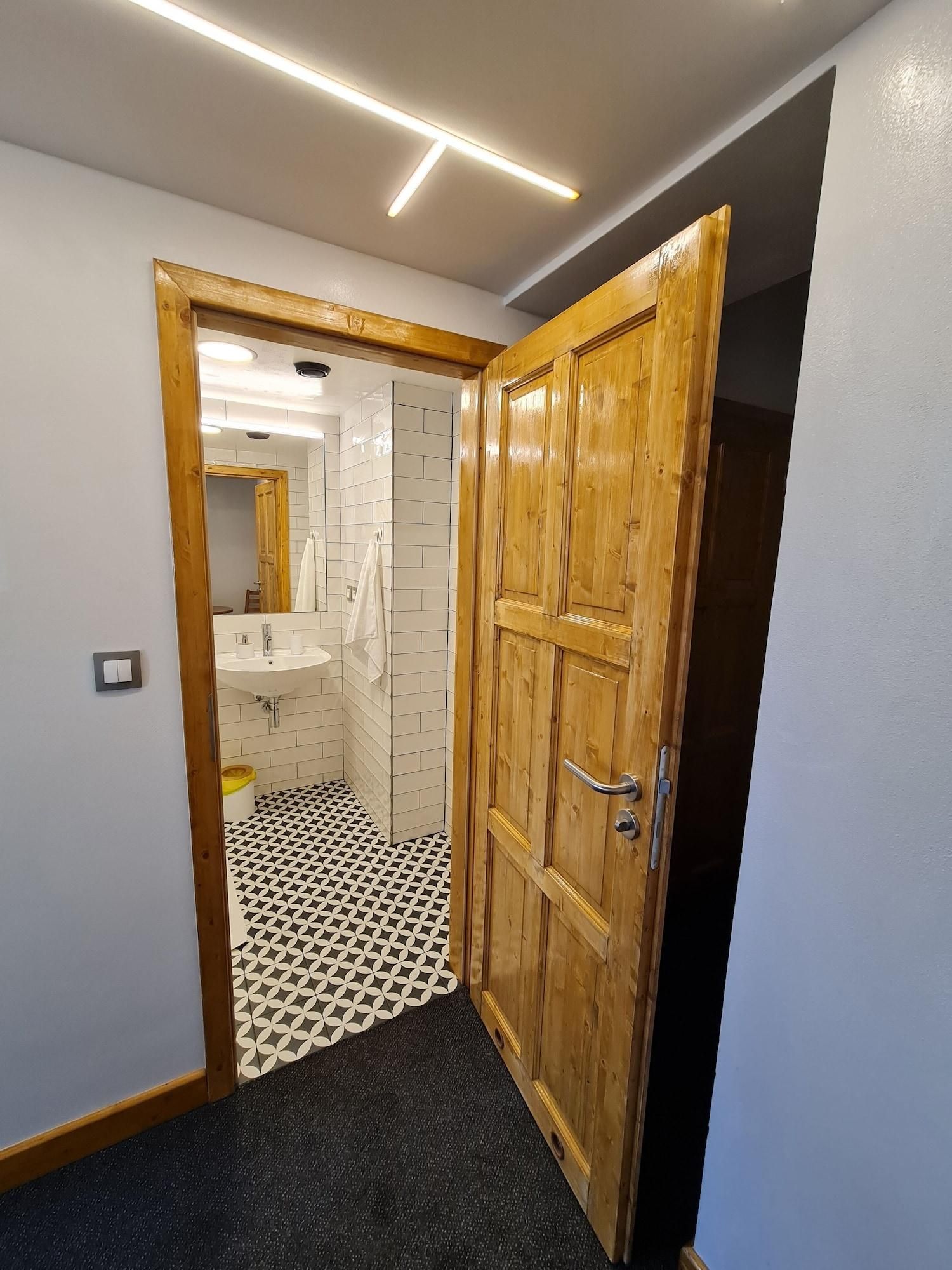 Een gang met een houten deur, een betegelde vloer en een badkamer door een geopende deur zichtbaar.