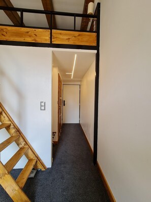 Un pasillo estrecho con una escalera de madera a un lado y una puerta al final.