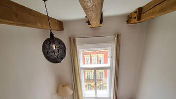 Een gezellige kamer met houten plafond, een bank met kussens, een klein houten tafeltje, een lamp en uitzicht op een gebouw door een raam.