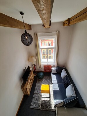Un cuarto acogedor con techo de madera, un sofá con cojines, una pequeña mesa de madera, una lámpara y vistas a un edificio a través de una ventana.