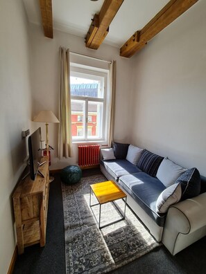 A modern living room with a sofa, a wooden side table, a lamp, and a view of a red building through a window. - Hotel S42 (Prague)