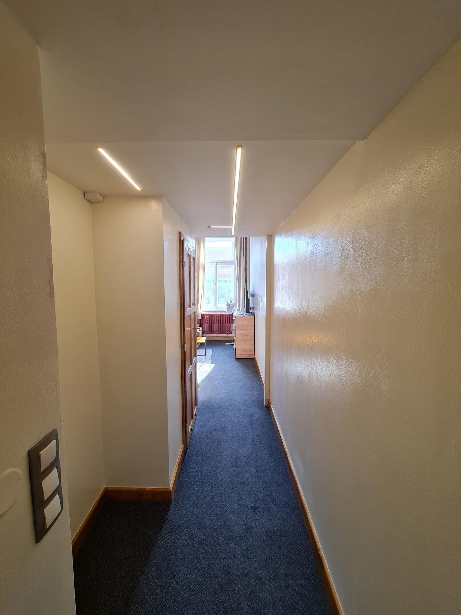 Un pasillo con pisos alfombrados, un radiador y una ventana con cortinas.