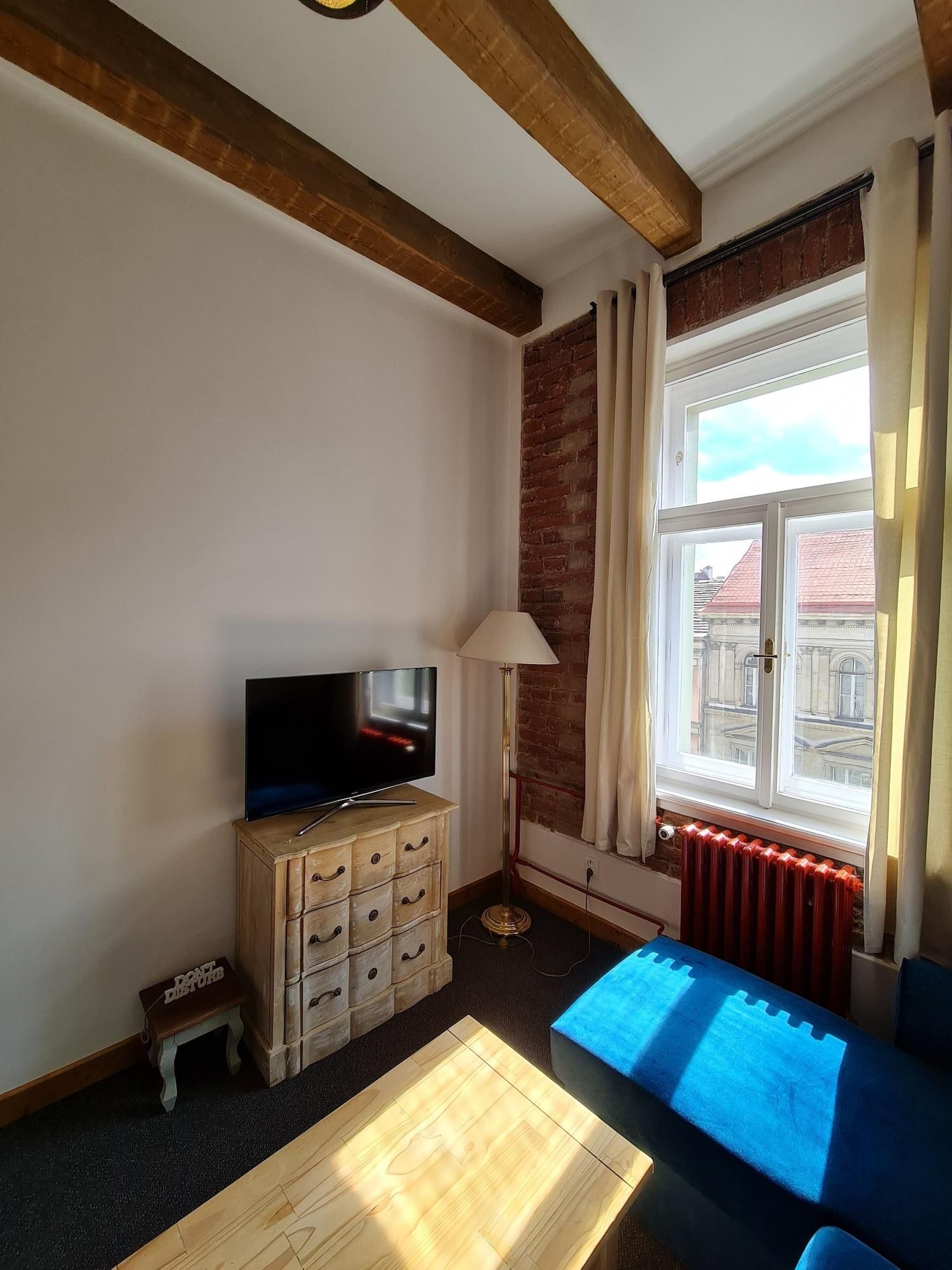 Una sala de estar con una televisión sobre un mueble de madera, una lámpara de pie, un sofá azul y una ventana con cortinas.
