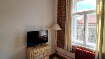 Una sala de estar con una televisión sobre un mueble de madera, una lámpara de pie, un sofá azul y una ventana con cortinas.