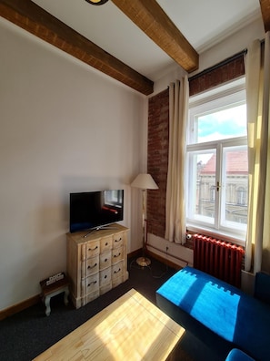 Un salon avec un téléviseur posé sur un meuble en bois, un lampadaire, un canapé bleu et une fenêtre avec des rideaux.