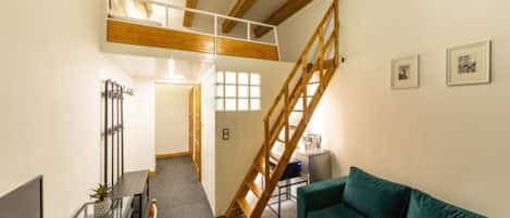 Un espacio habitable tipo loft con techo alto, vigas de madera a la vista, un sofá verde, una mesa de comedor y una escalera.