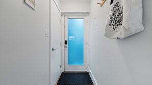 A hallway with a white door, a black doormat, and a white wall with a hanging bag. - Modern, Quiet, Fully Furnished Suite in the Heart of Lower Lonsdale (North Vancouver)