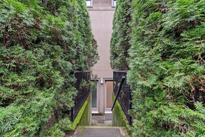 Un étroit sentier bordé de grands arbres à feuilles persistantes verdoyants mène à l’entrée d’un bâtiment, qui est protégée par un portail en métal noir.