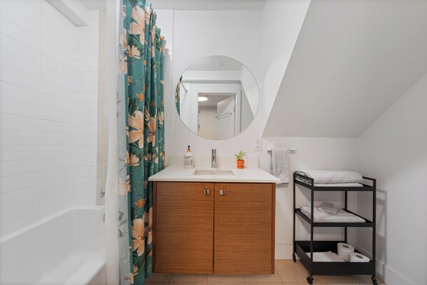 Une salle de bain avec un meuble-lavabo en bois, un évier, un miroir, un rideau de douche à motifs floraux et une étagère noire avec des serviettes.